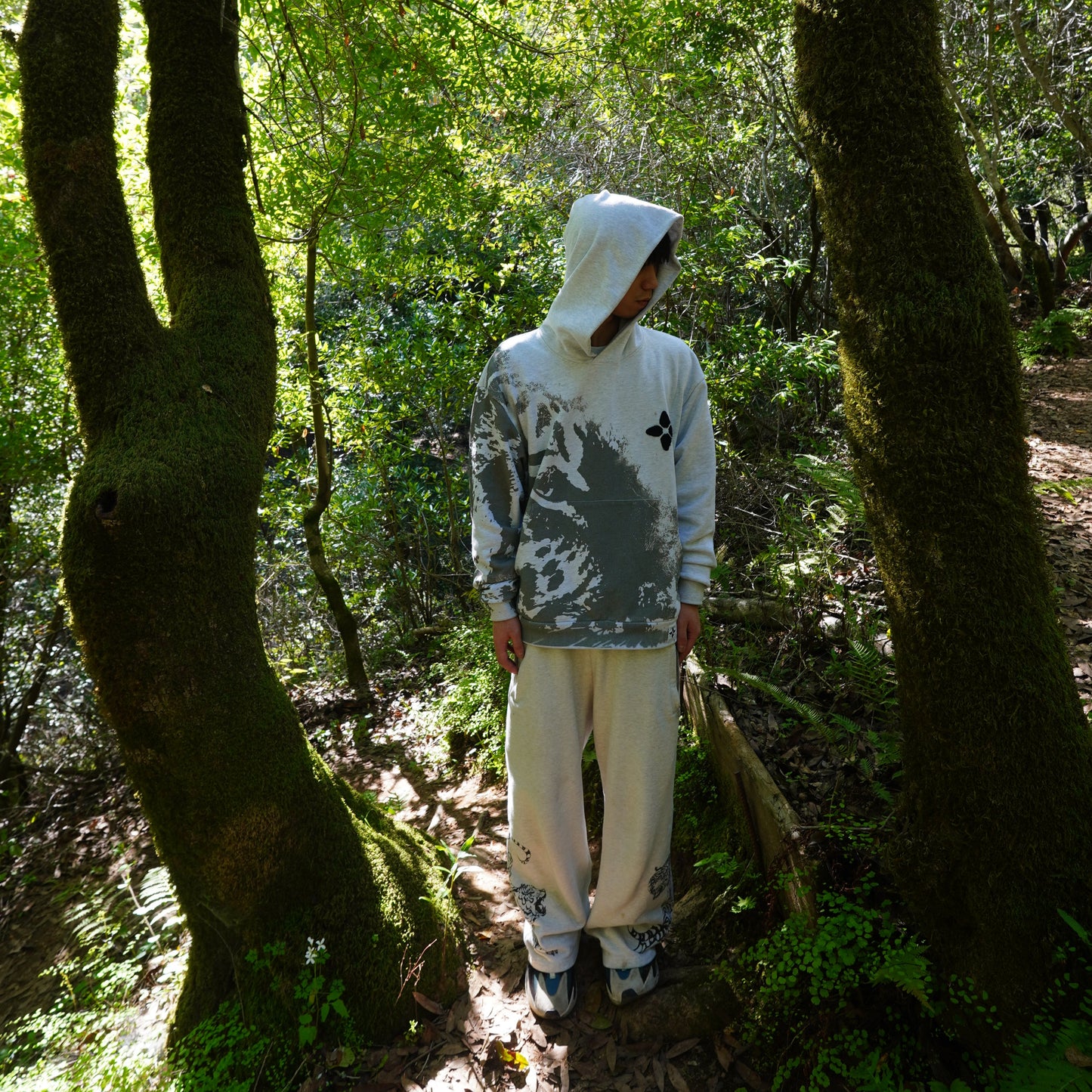 Person wearing a white shirt with black text and graphics standing between two trees in a forest.